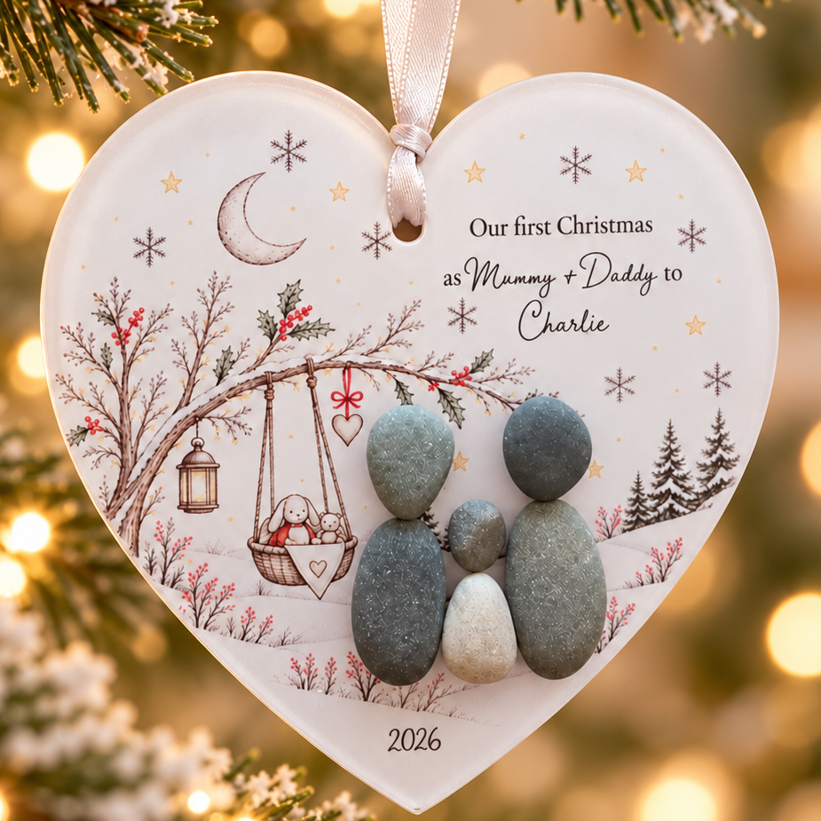 Close-up of the Our First Christmas decoration: two pebble parents and a baby pebble beside a swinging cradle, dated 2026
