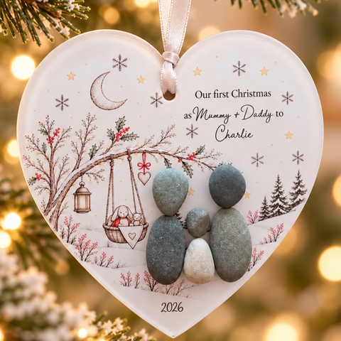 Close-up of the Our First Christmas decoration: two pebble parents and a baby pebble beside a swinging cradle, dated 2026