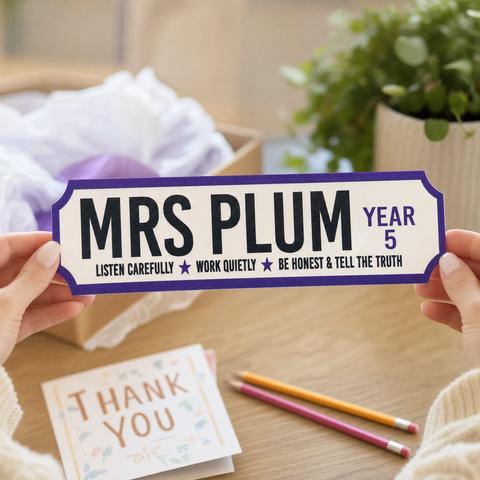 Person holding a nameplate with 'MRS PLUM YEAR 5' on a wooden table with stationery items.