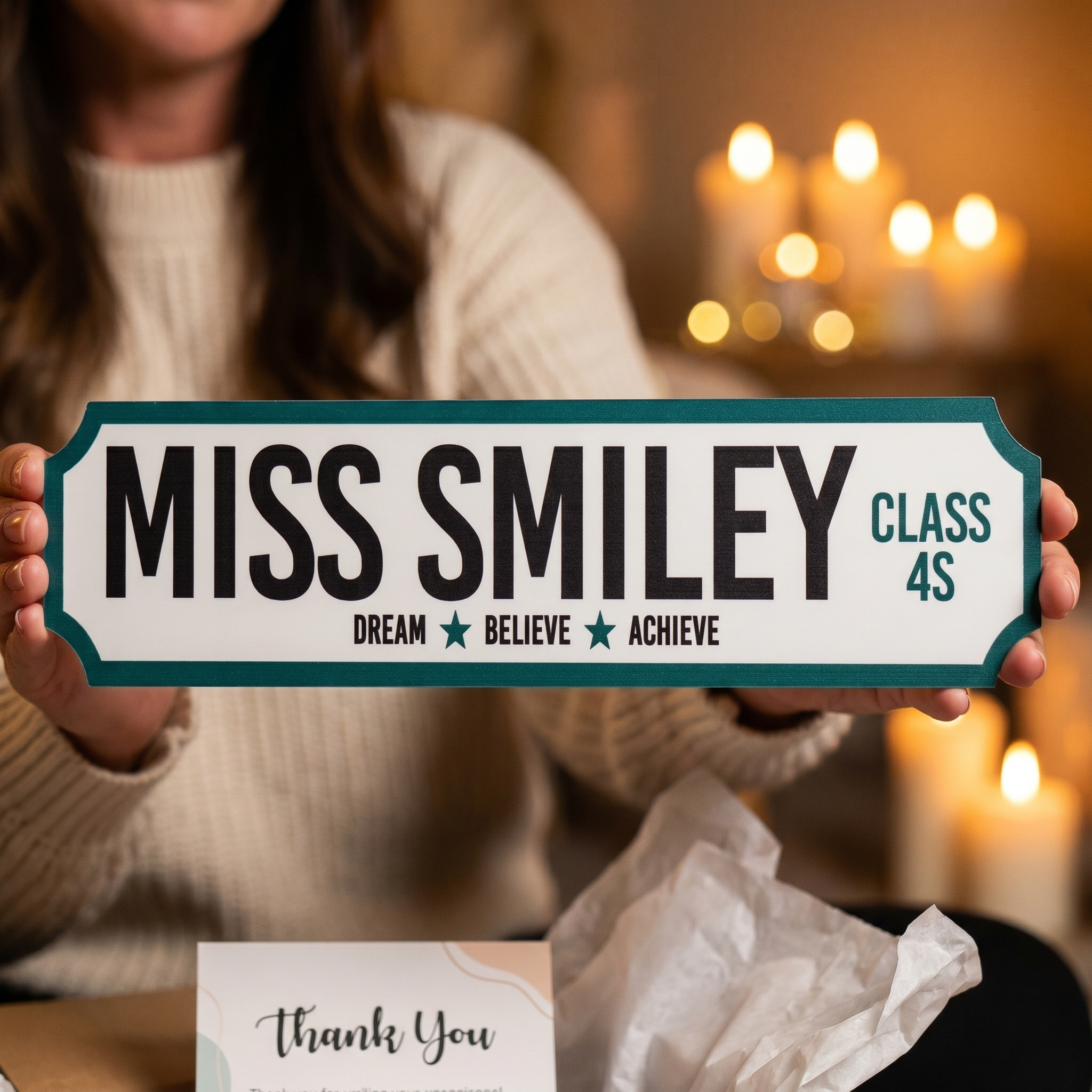 Person holding a sign that reads 'MISS SMILEY CLASS 4S' with a blurred candlelit background.