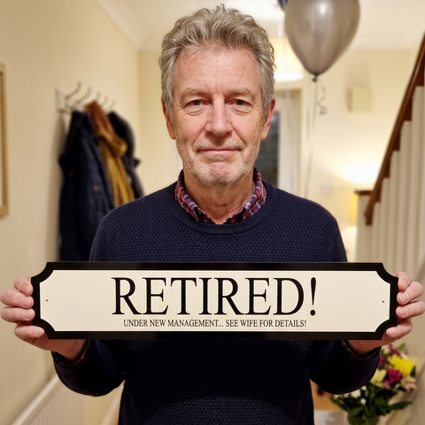 A man standing indoors holding a decorative sign that reads 'RETIRÉ!' with smaller text underneath. Balloons are visible in the background near a staircase.